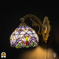 Verlichte Tiffany wandlamp Royal Mosaic met lavendelblauwe glas-in-lood kap, crèmegele bloemmotieven en kleurrijke juwelen op bronzen sierlijke arm