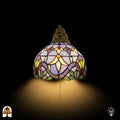 Frontaal aanzicht verlichte Tiffany wandlamp Royal Mosaic, koepelkap in lavendel met crèmegele bloemen en groene bladrand, trekschakelaar zichtbaar