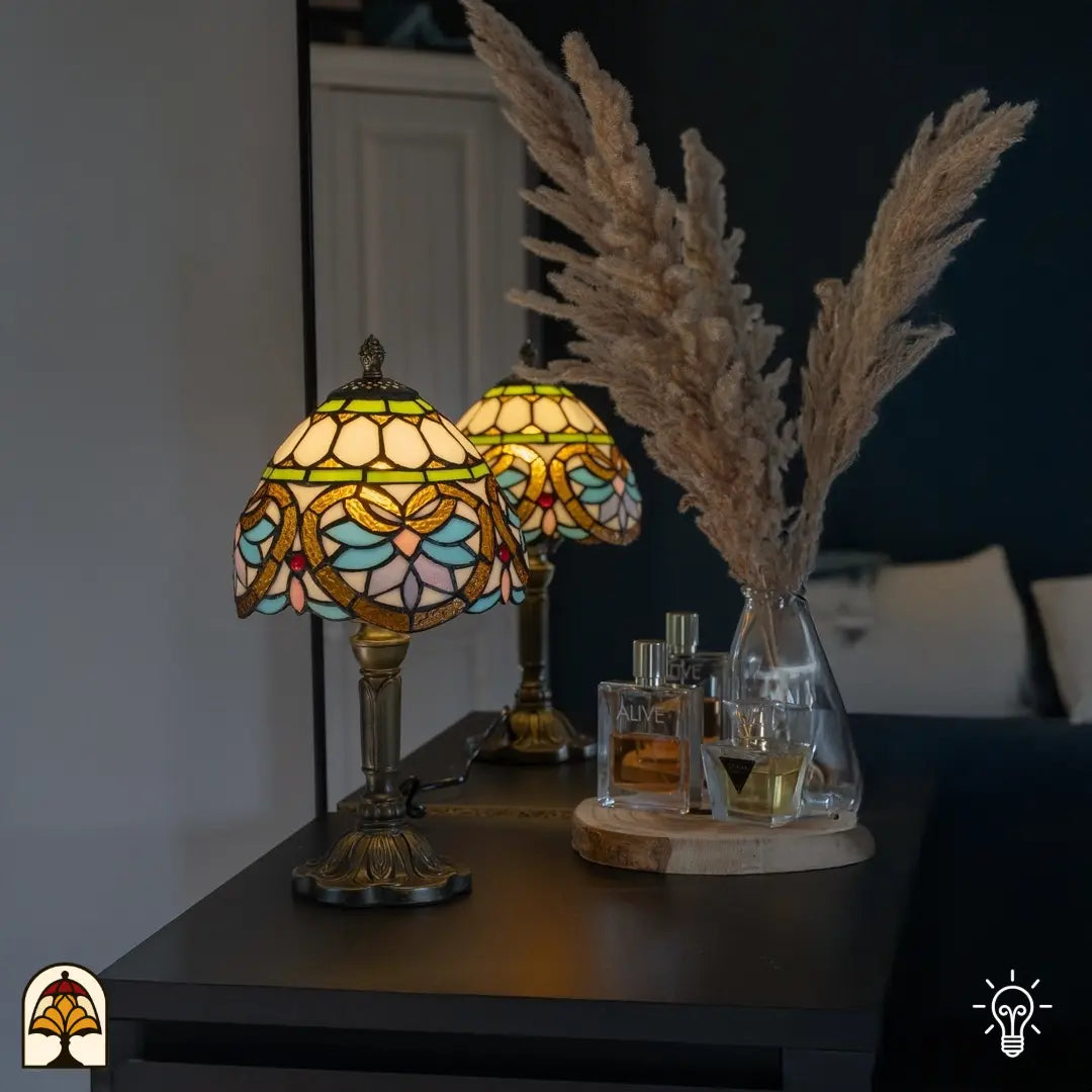 Lampe de table Tiffany à motif floral dans des tons pastel doux, posée sur un meuble sombre à côté d'un vase en verre avec herbe de la pampa et flacons de parfum, diffuse une lumière chaude dans une chambre chaleureuse.