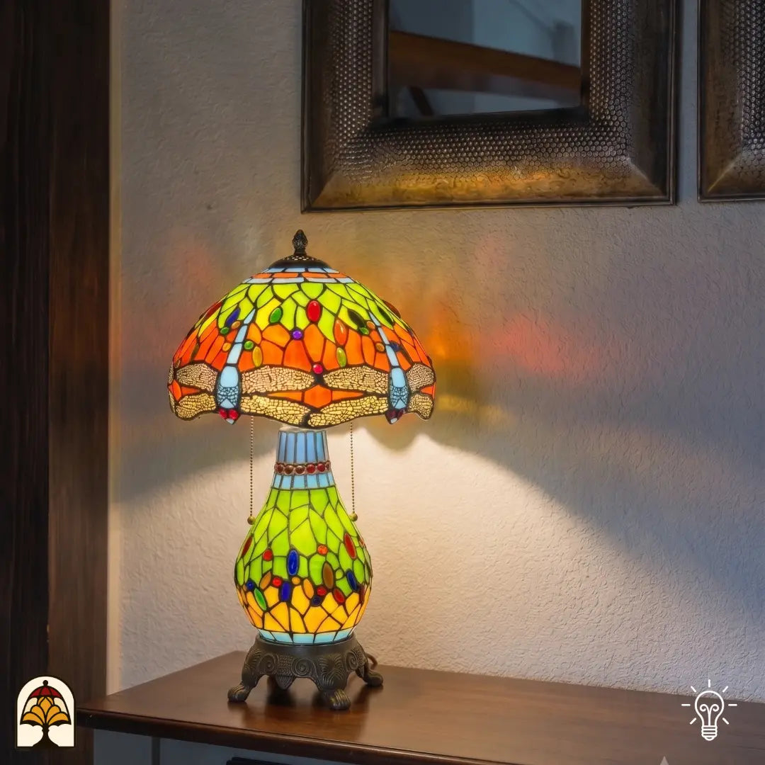 Lampe de table Tiffany Libellula Lumina ⌀30 cm allumée dans un intérieur chaleureux – abat-jour en vitrail orange et jaune-vert à motif de libellule, pied en bronze à pattes de lion, lumière chaude