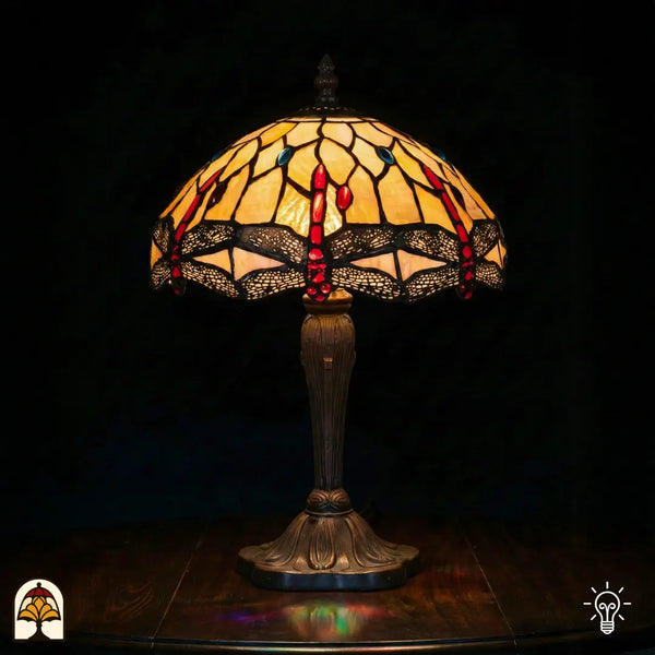 Lampe de table Tiffany Libelle Elegance allumée avec abat-jour en dôme jaune ambré, accents rouges en forme de goutte et accents ronds bleus en vitrail, motifs de libellules le long du bord, pied vase en bronze cannelé, fond sombre