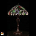 Onverlichte Tiffany tafellamp Sylva Berria op donkere achtergrond, kap met lila en rode druiventrosjes en groene bladeren, slanke bronzen Art Nouveau vaasvoet