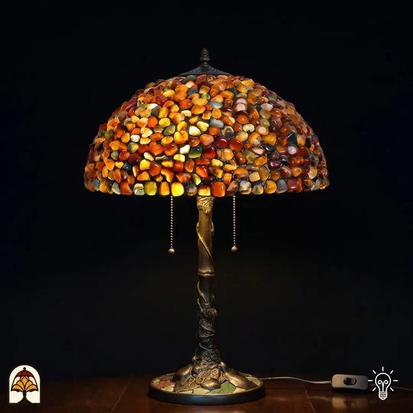 Lampe de table Tiffany Stone Mosaic Amber ⌀40 cm allumée sur fond sombre – abat-jour en dôme en pierres polies ambrées, orange et ocre, pied en forme de tronc d'arbre en bronze avec deux interrupteurs à chaînette