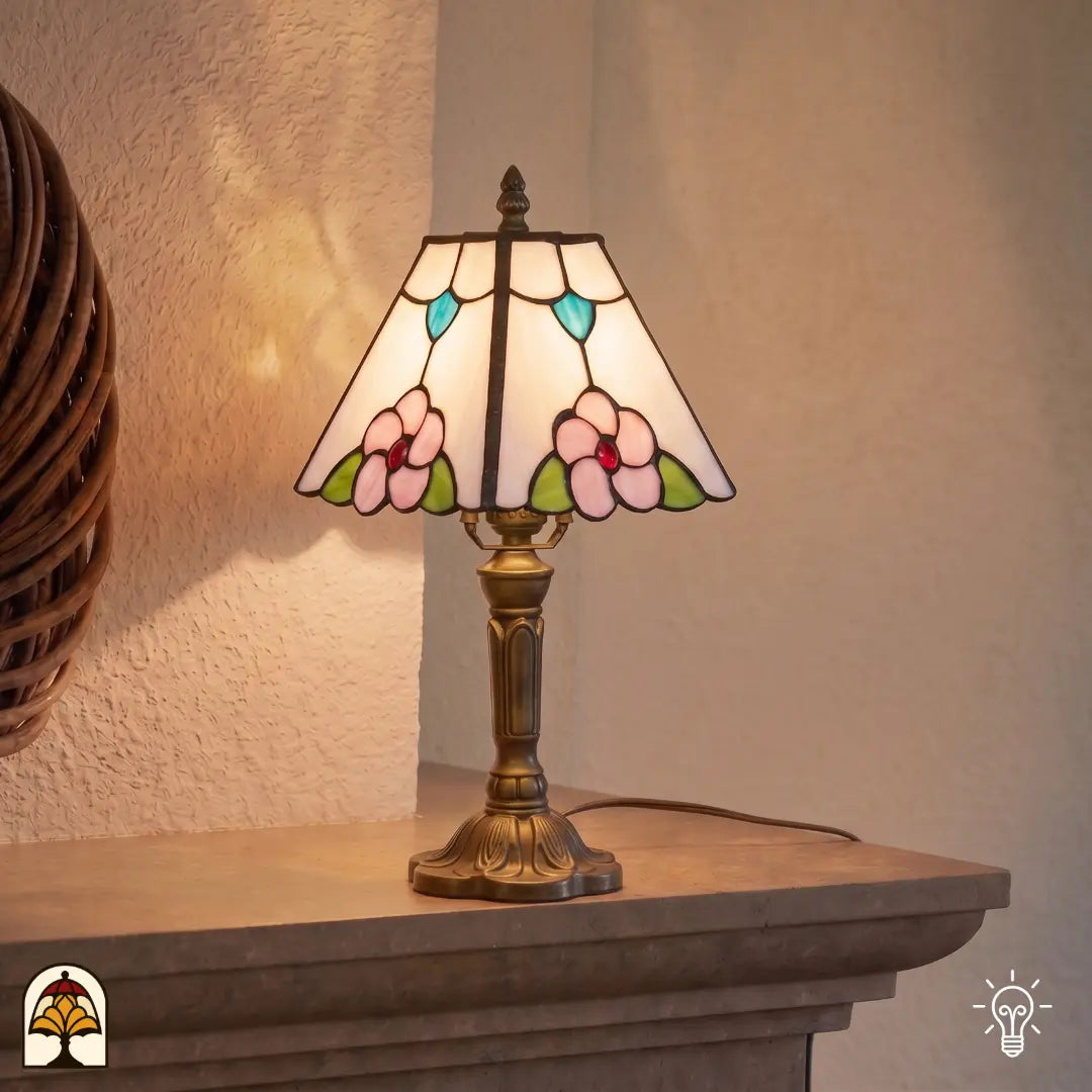 Lampe de table Tiffany Rosa Zaffiro ⌀20 cm allumée dans un intérieur chaleureux sur une table en bois – abat-jour crème avec fleurs roses et accents de feuilles bleues, pied en bronze, lumière chaude