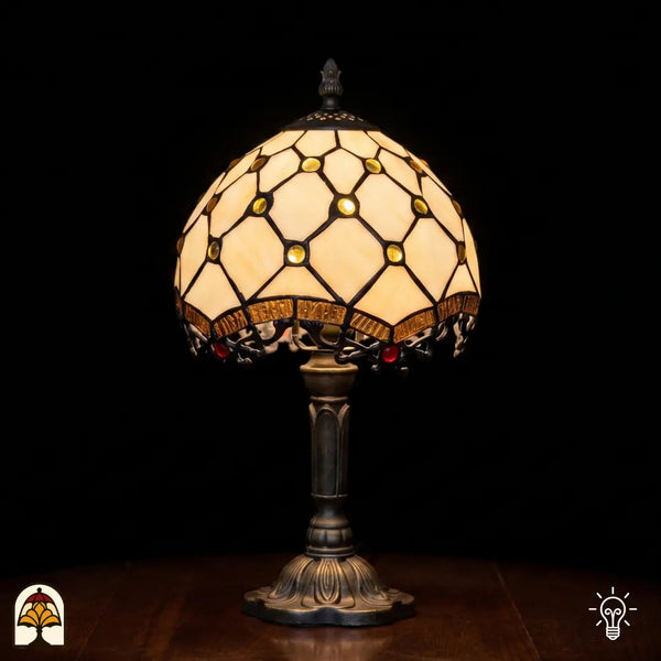 Lampe de table Tiffany Reticolo d'Oro ⌀20 cm allumée sur fond noir – abat-jour crème avec cabochons en verre dorés et motif losange, pied en bronze