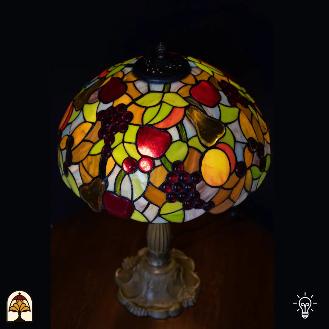 Gros plan abat-jour allumé lampe de table Tiffany Frutta Viva vue de dessus — raisins, pommes rouges et poire jaune en verre, feuilles vertes, aplats orange/jaunes