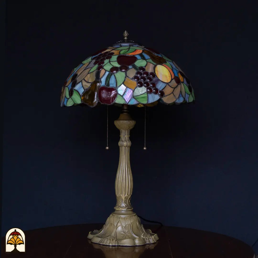 Lampe de table Tiffany Frutta Viva éteinte sur fond sombre — abat-jour avec raisins, pommes et poire, surfaces bleues/vertes, pied colonne en bronze