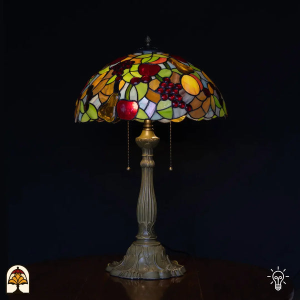 Lampe de table Tiffany Frutta Viva allumée sur fond noir — abat-jour dôme avec pommes rouges, raisins violets et poire jaune en verre, feuilles vertes, pied colonne en bronze avec interrupteurs à chaînette
