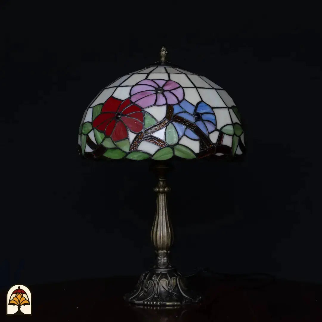 Lampe de table Tiffany Aurora Bloom éteinte sur fond noir — abat-jour blanc avec fleurs roses, rouges et bleues, branches dorées, feuilles vertes, pied colonne en bronze
