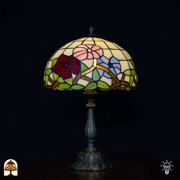 Lampe de table Tiffany Aurora Bloom allumée sur fond noir — abat-jour dôme crème avec fleurs roses, rouge foncé et bleues sur branches dorées, feuilles vertes, pied colonne en bronze