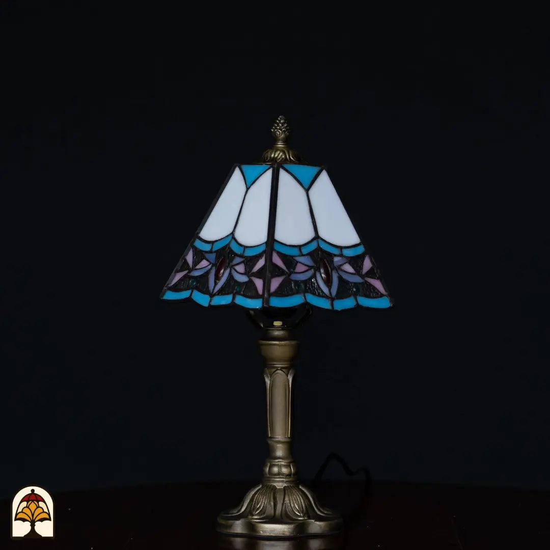 Lampe de table Tiffany éteinte sur fond sombre — abat-jour blanc avec bords bleus, fleurs de lotus violettes et pierres précieuses rouges, pied chandelier classique