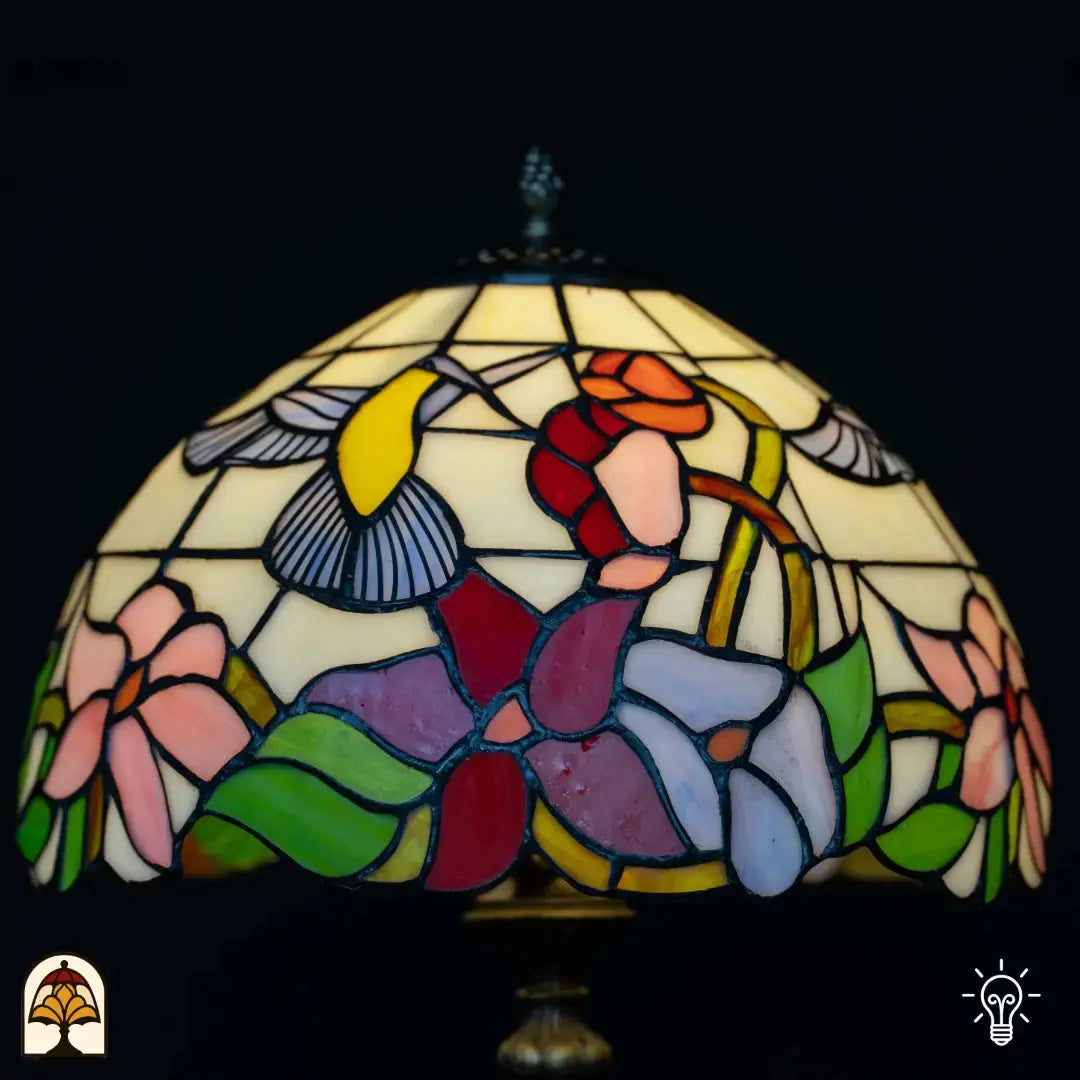 Gros plan abat-jour allumé lampe de table Tiffany Colibrì — verre crème avec colibri (ailes jaunes/bleues), fleurs rouges et orange, fleurs violettes et bleues en bas, feuilles vertes