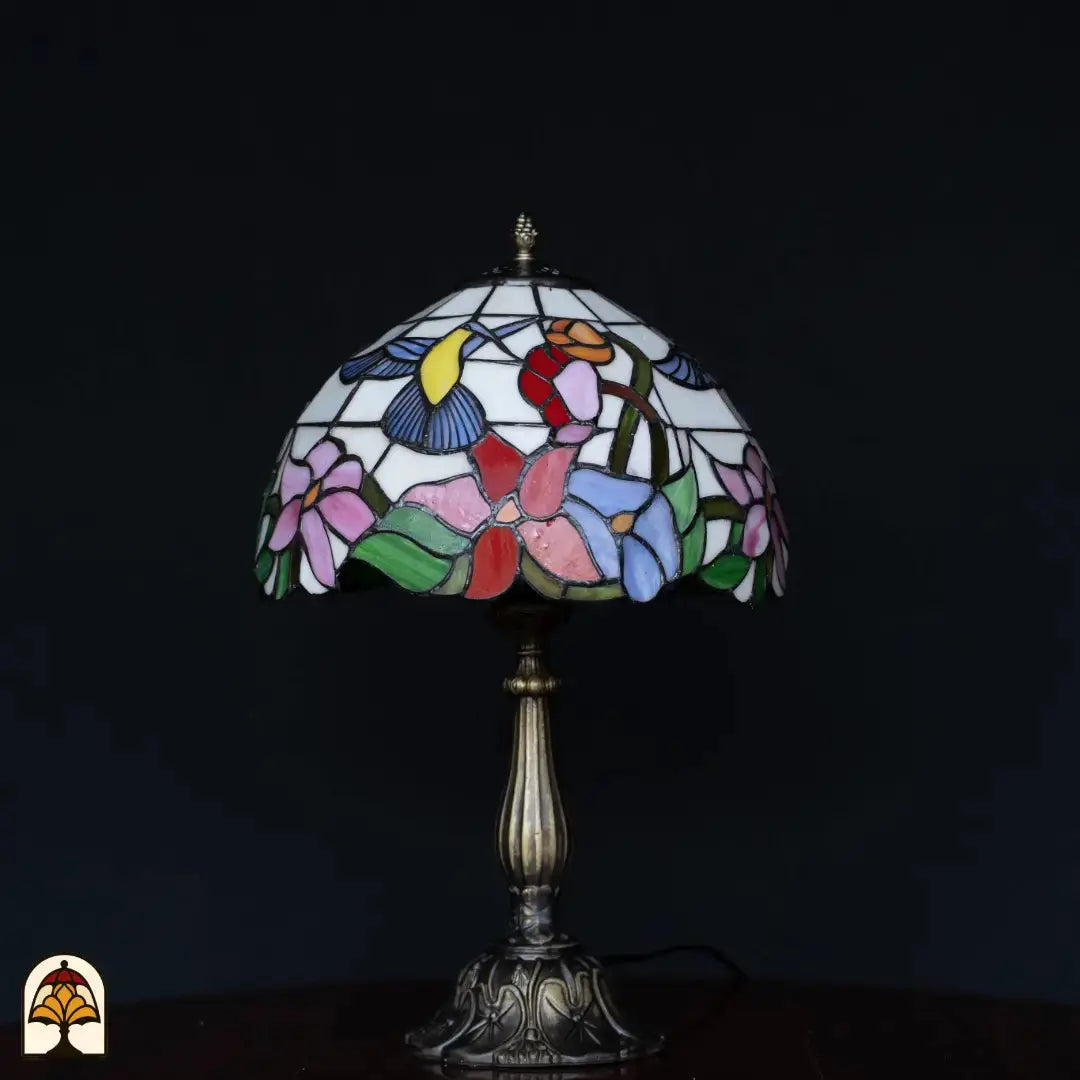 Lampe de table Tiffany Colibrì éteinte sur fond noir — abat-jour blanc/crème avec colibri et fleurs en rose, rouge, bleu et vert, pied colonne en bronze