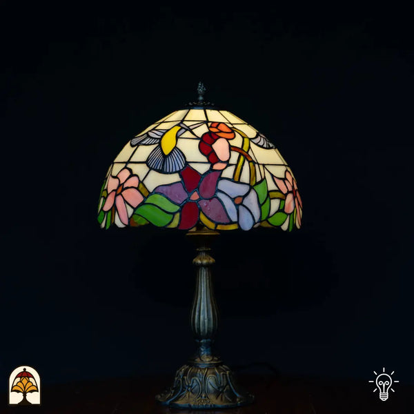 Lampe de table Tiffany Colibrì allumée sur fond noir — abat-jour dôme crème avec colibri et fleurs colorées en rose, rouge, violet et bleu, feuilles vertes, pied colonne en bronze
