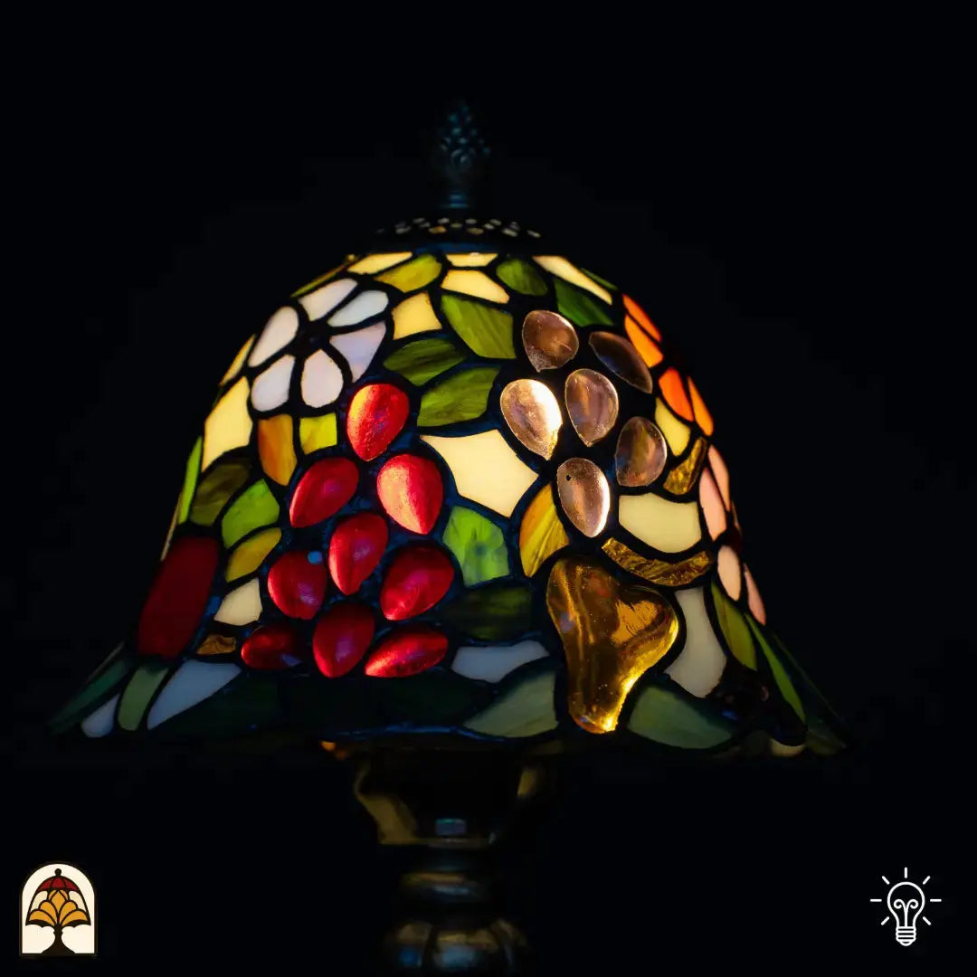 Gros plan abat-jour Tiffany allumé — raisins rouges, fruits ambrés/dorés en forme de poire, fleurs blanches et feuilles vertes sur fond jaune