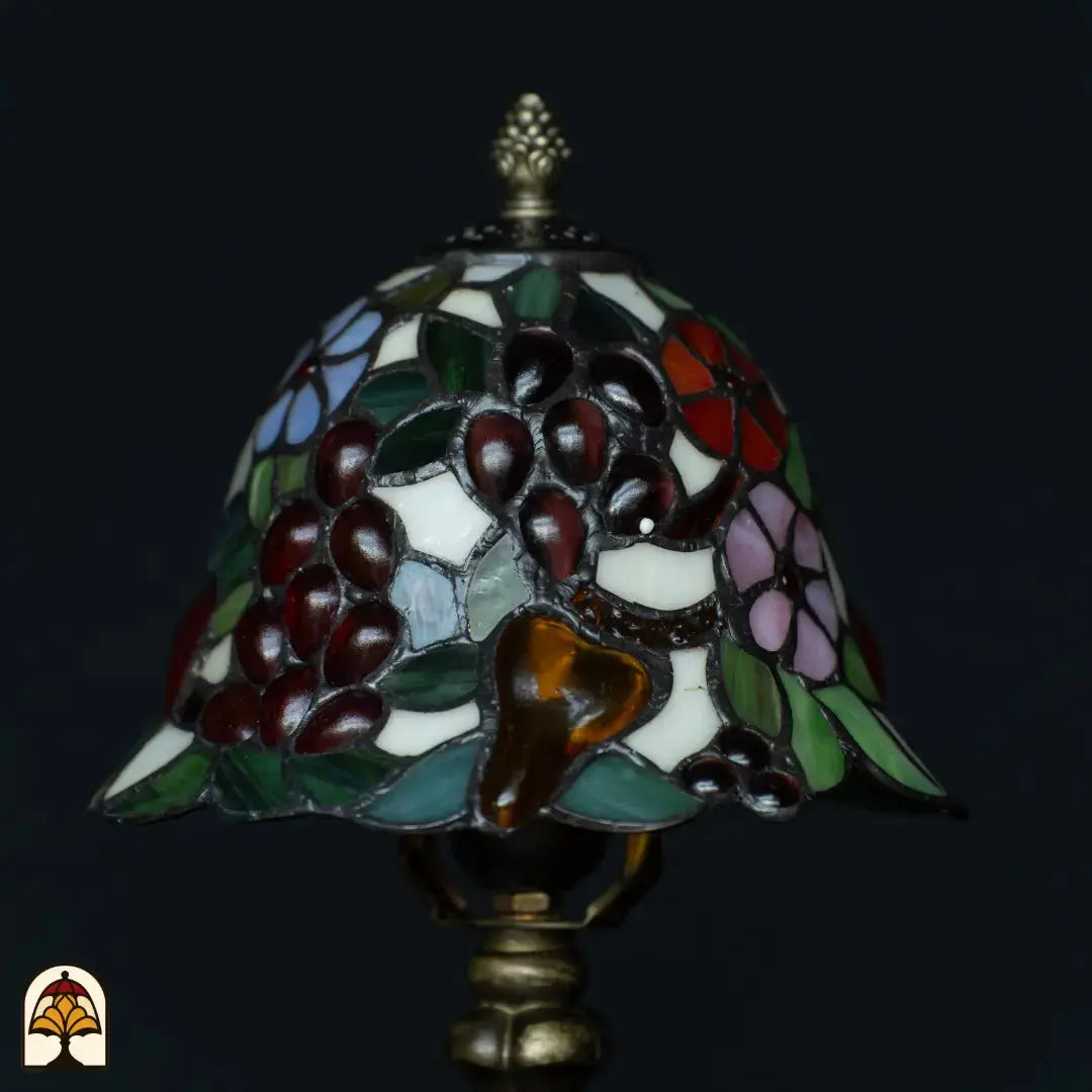 Gros plan abat-jour Tiffany éteint — raisins bordeaux, fruit ambré en forme de poire, feuilles vertes, fleurs roses et lilas sur fond blanc