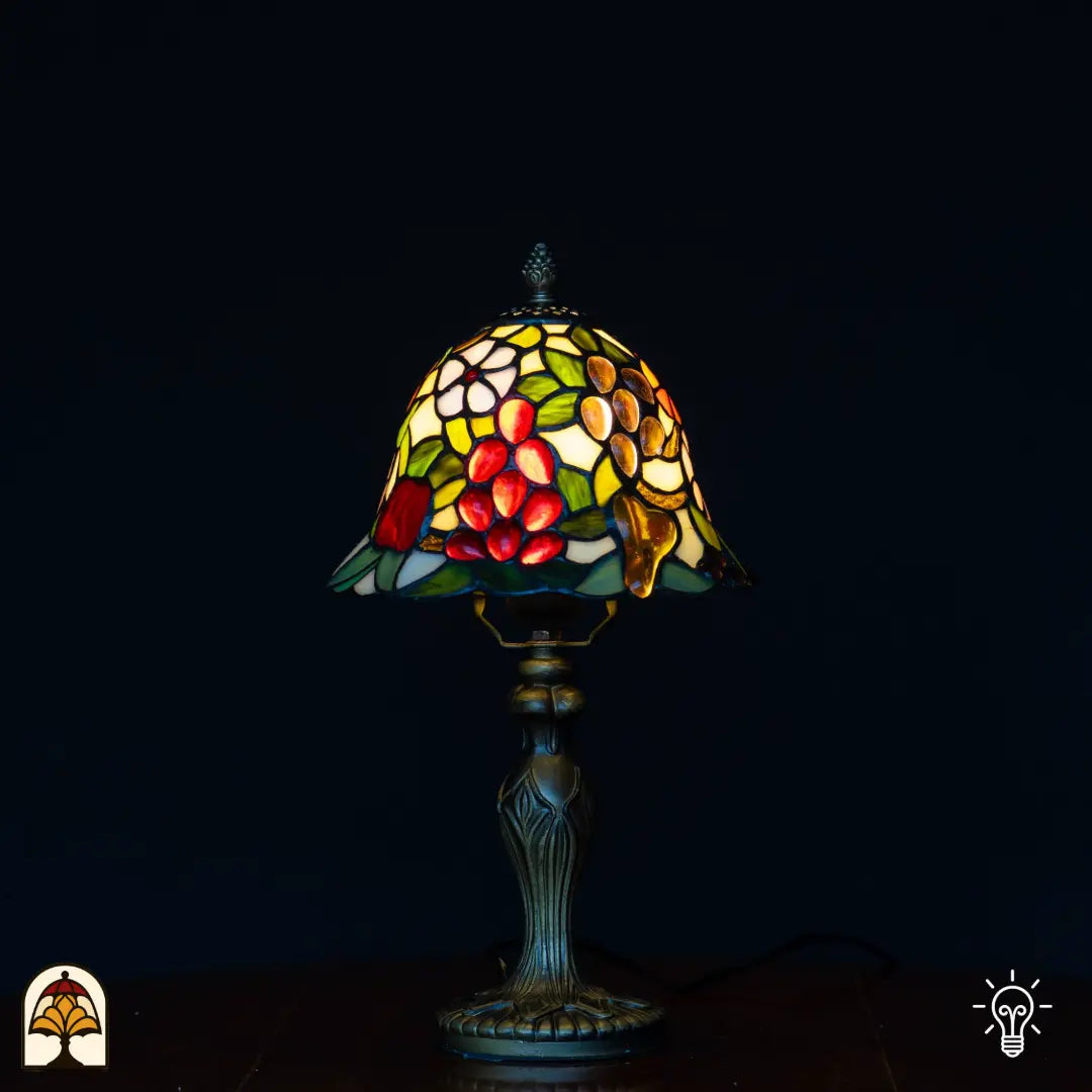 Lampe de table Tiffany allumée avec abat-jour en forme de cloche à raisins rouges, fruits en forme de poire ambrés/dorés, fleurs blanches et feuilles vertes sur fond jaune, pied chandelier classique, fond noir