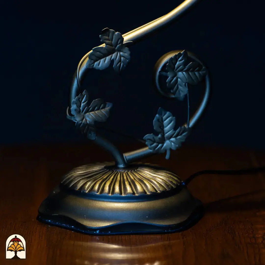 Gros plan pied — bras courbé sombre avec décorations en forme de feuilles, base ronde ovale avec motif nervuré doré/bronze, câble d'alimentation visible