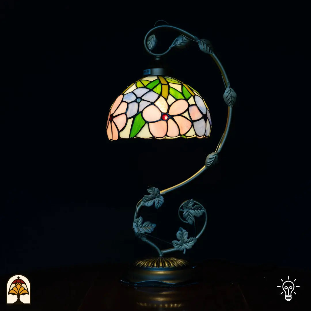 Lampe de table Tiffany allumée avec bras courbé en col de cygne, abat-jour en dôme avec fleurs roses et feuilles vertes sur fond crème, pied sombre en forme de feuille avec base ronde, fond noir