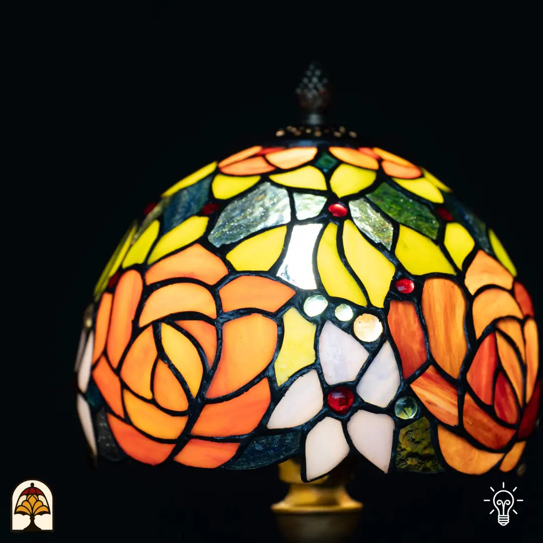 Gros plan abat-jour allumé lampe de table Tiffany Fiore — roses orange/rouges dominantes, feuilles jaunes et vertes tout autour, cabochons en verre rouges et verts, petites fleurs blanches