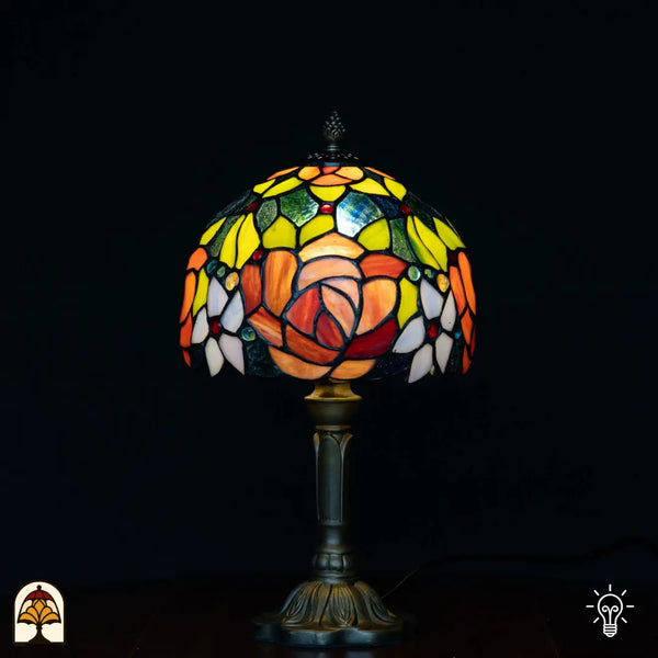 Lampe de table Tiffany Fiore allumée sur fond noir — abat-jour dôme avec grandes roses orange, feuilles jaunes/vertes, cabochons en verre rouges et bleus, pied colonne en bronze
