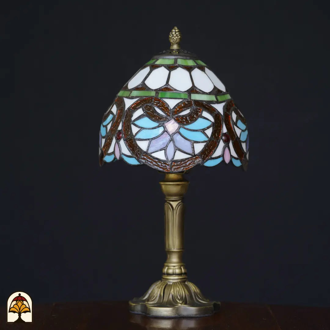 Lampe de table Tiffany Lucette éteinte sur fond noir – abat-jour crème avec bande verte, formes de feuilles baroques brunes, pétales bleu clair et roses, pierres précieuses rouges, pied colonne en bronze