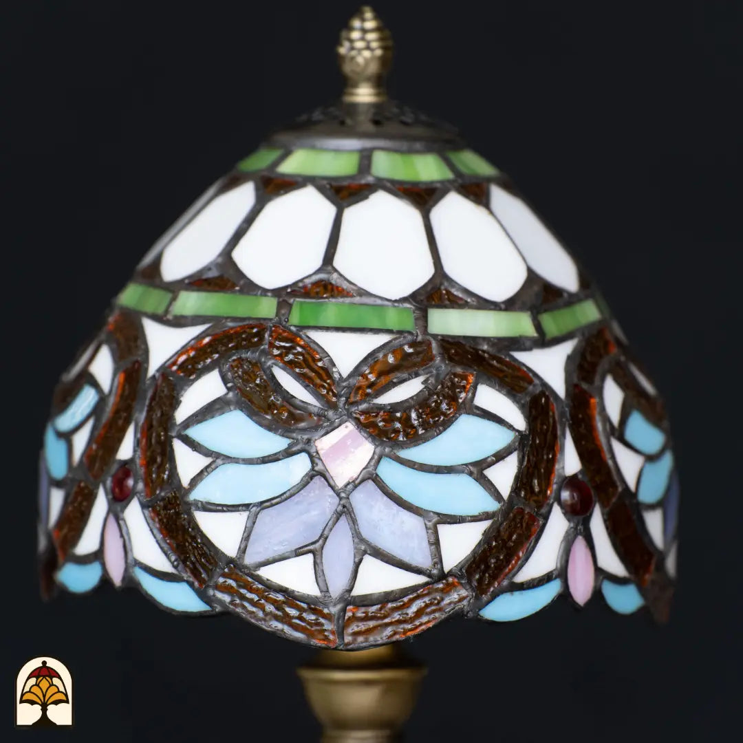 Gros plan abat-jour dôme éteint lampe de table Tiffany Lucette – verre blanc avec bandeau décoratif vert, formes de feuilles brunes, pétales bleu clair et roses et cabochon rouge