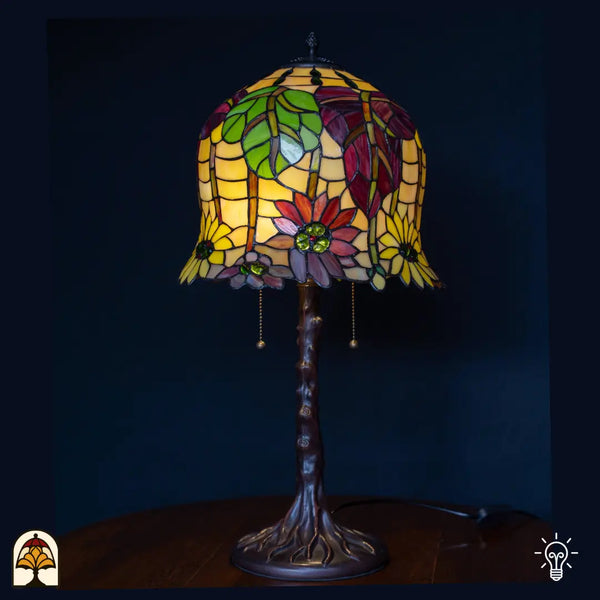 Lampe de table allumée avec haut abat-jour Tiffany en forme de cloche ambré/jaune à fleurs rouges et violettes et feuilles vertes, pied en forme de tronc d'arbre avec interrupteurs à chaînette, fond bleu foncé