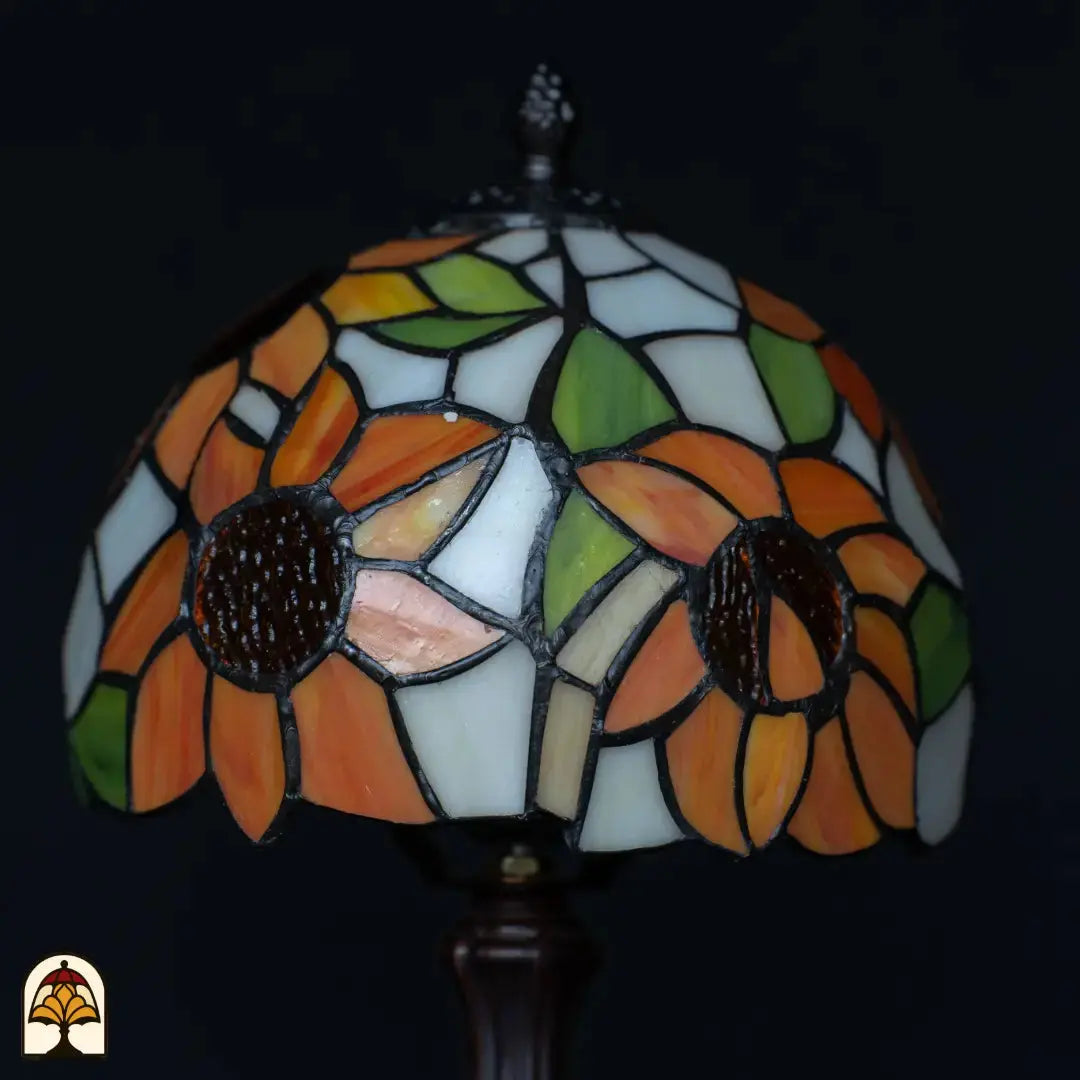 Gros plan abat-jour lampe de table Tiffany Solea éteinte — pétales de tournesol orange à cœur brun foncé, surfaces crème/blanches et bleues, feuilles vertes