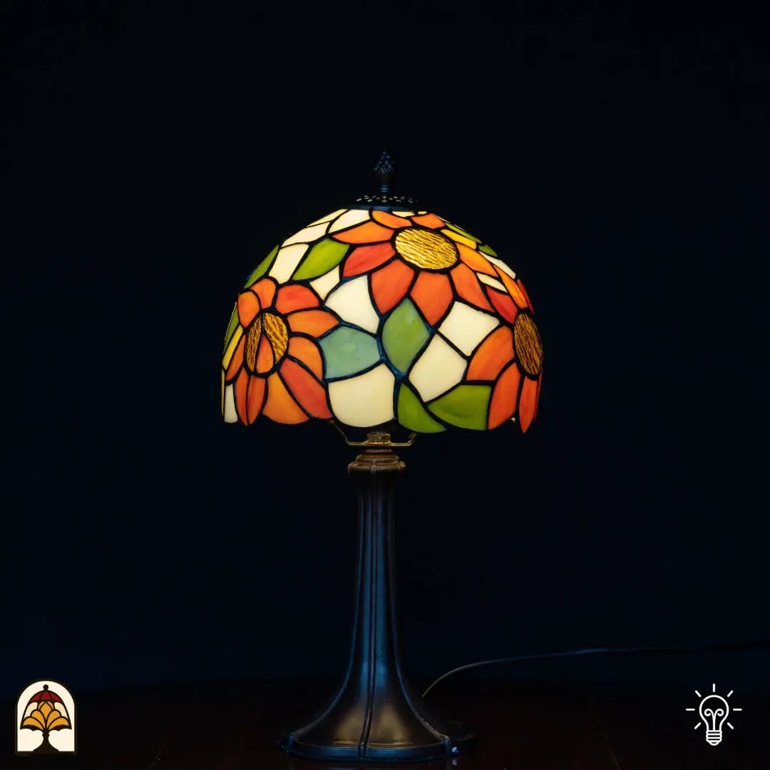 Lampe de table Tiffany Solea allumée sur fond noir — abat-jour dôme crème/blanc avec tournesols orange et coeurs jaunes, feuilles vertes, pied Art Nouveau élancé en bronze