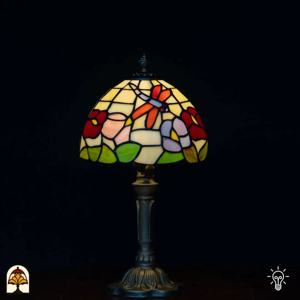 Lampe de table Tiffany allumée avec abat-jour en dôme à libellule orange/rouge, fleurs rouges, fleurs roses et violettes et feuilles vertes sur fond crème/jaune, pied chandelier classique, fond noir