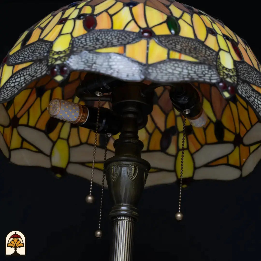 Gros plan douille lampadaire Tiffany Libellula Aurea éteint — deux ampoules LED dans douille en bronze, interrupteurs à chaînette, abat-jour coloré à bordure de libellules en arrière-plan