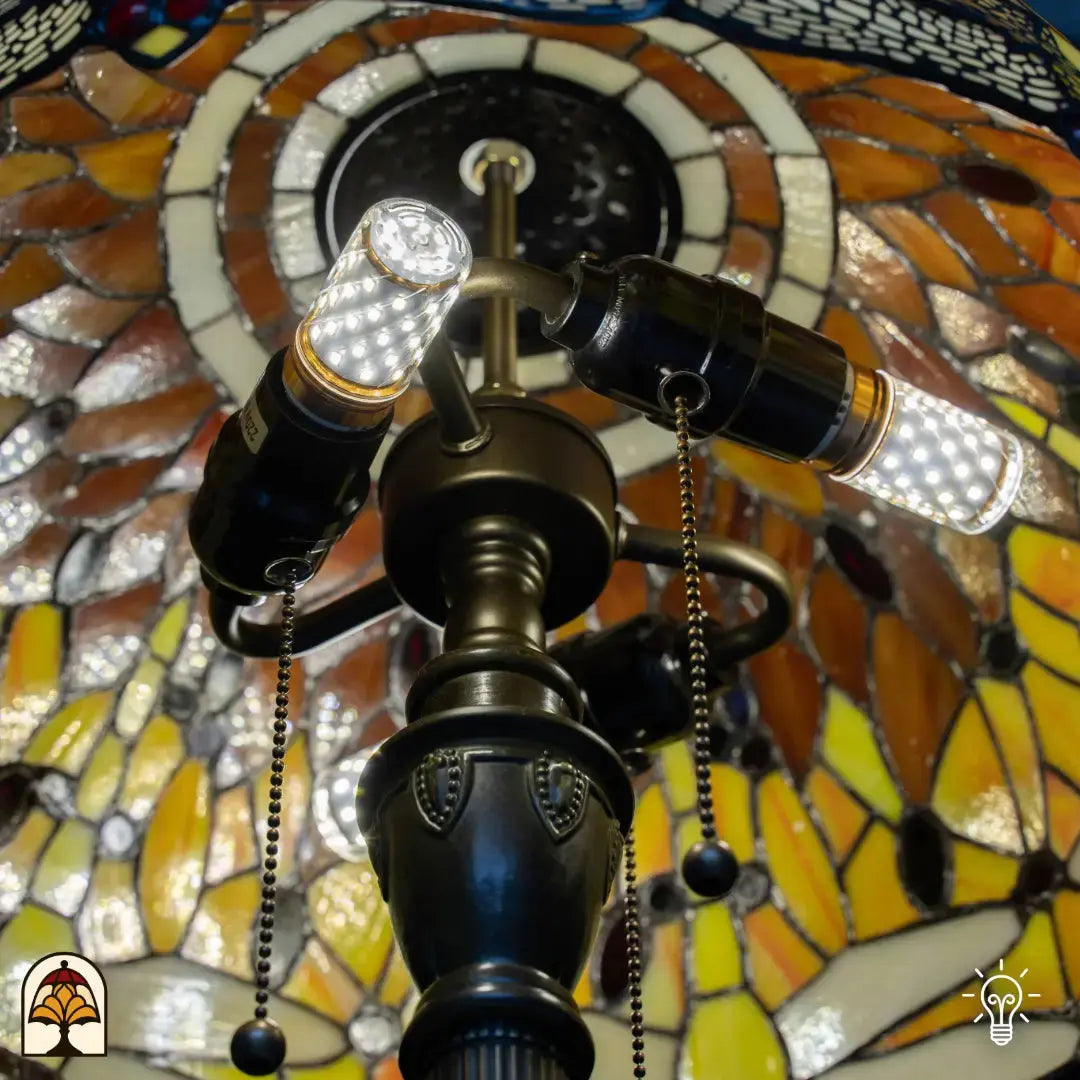 Gros plan de la douille allumée du lampadaire Tiffany Libellula Aurea vue de dessous — deux ampoules LED allumées dans une double douille en bronze, interrupteurs à chaînette, verre ambré/jaune allumé tout autour