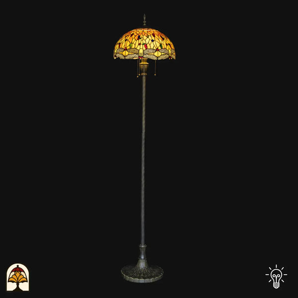 Lampadaire Tiffany Libellula Aurea allumé sur fond noir — abat-jour dôme ambré/jaune avec bordure à libellules, long fût avec base large en bronze, interrupteurs à chaînette